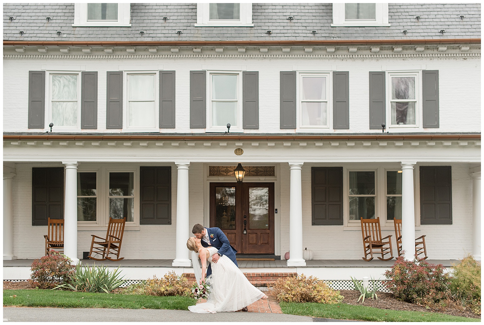 groom dipping his bride back as they kiss in front of white and gray farmhouse at historic ashland