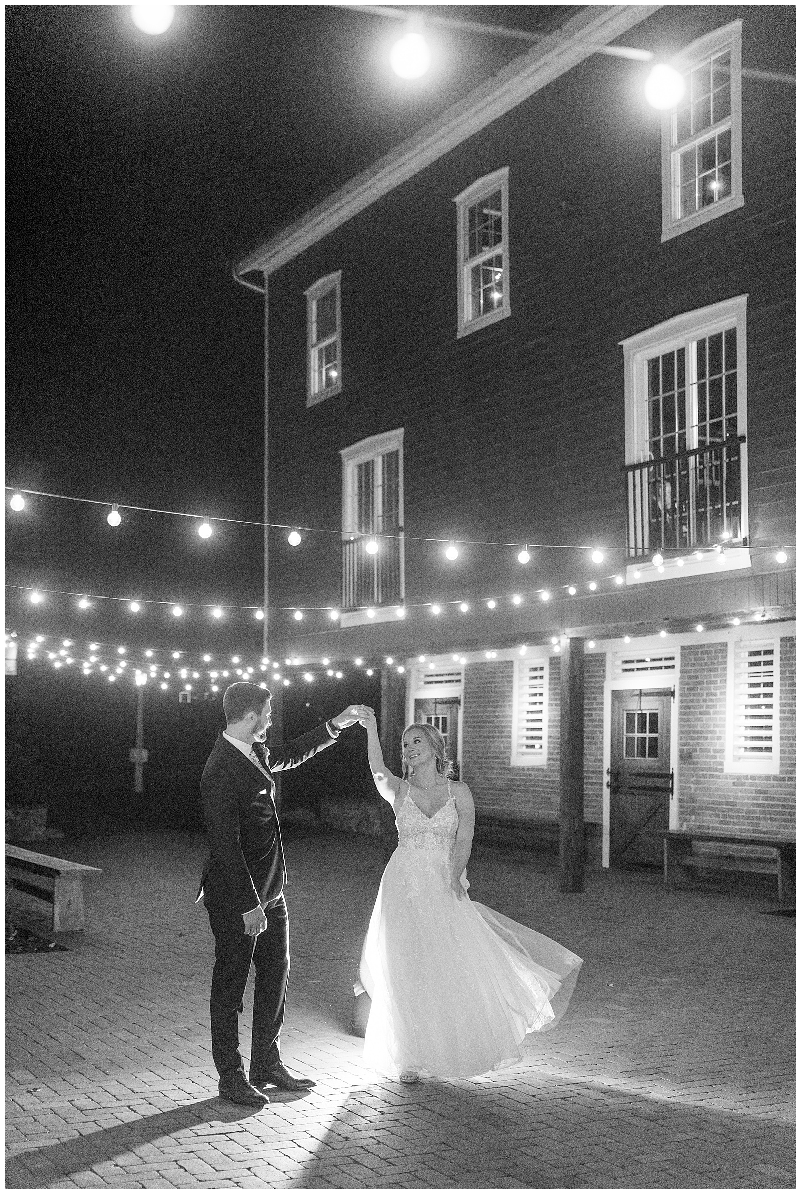 black and white photo of groom twirling his bride under rows of glowing string lights outside barn at historic ashland