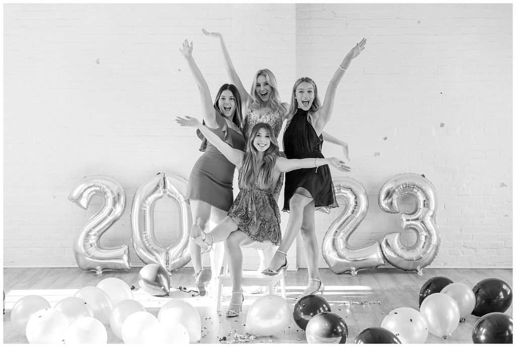 black and white photo of four class of 2023 girls crowded together with excitement by white brick wall in lancaster pennsylvania