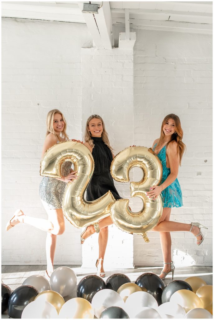 three senior spokesmodels wearing party dresses and holding large "2" and "3" balloons at the white room