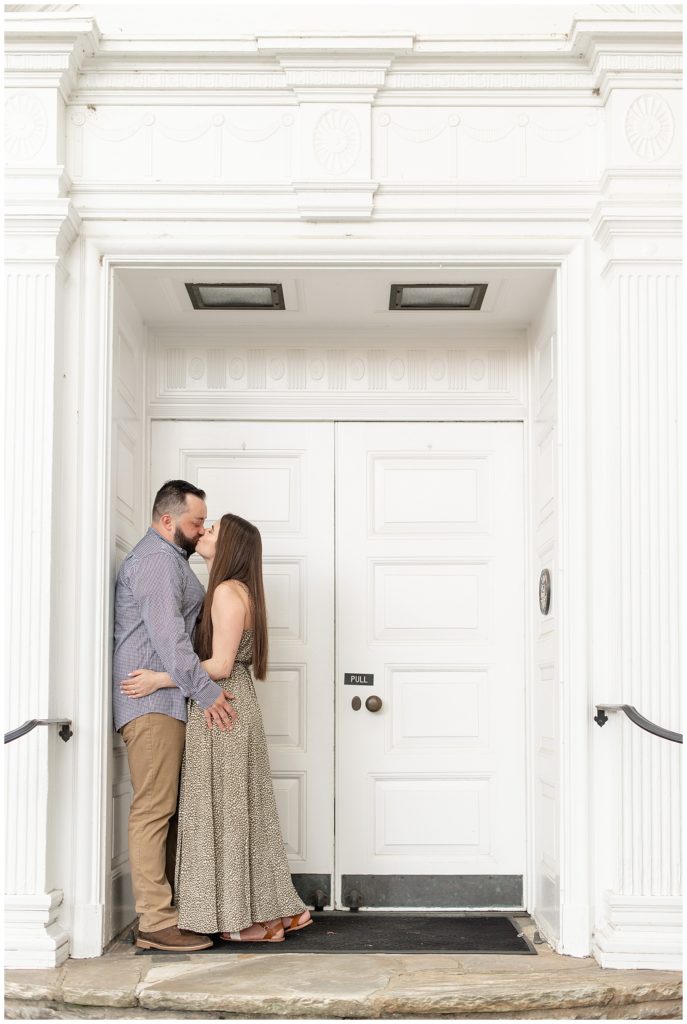 engaged couple kissing as guy leans against white door frame of historic building in downtown lititz pennsylvania