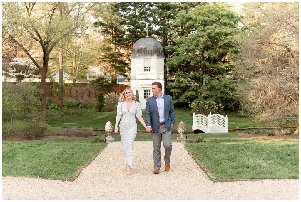 engaged couple holding hands and walking towards camera in manicured garden with small white tower behind them in annapolis maryland
