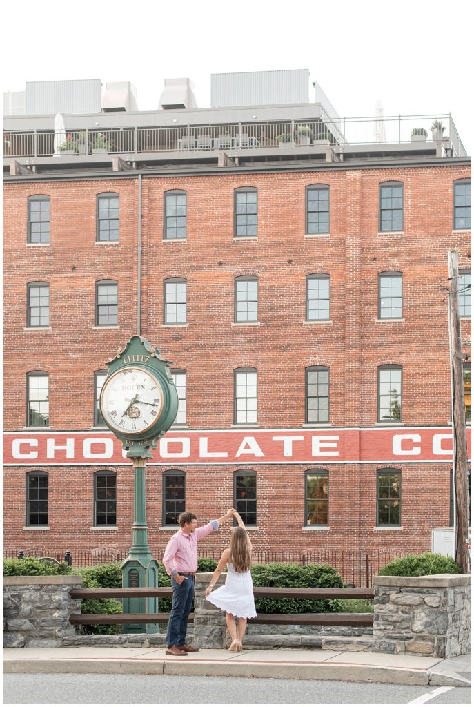 guy twirling girl under his left hand by old clock and historic brick wilbur chocolate building in downtown lititz pennsylvania