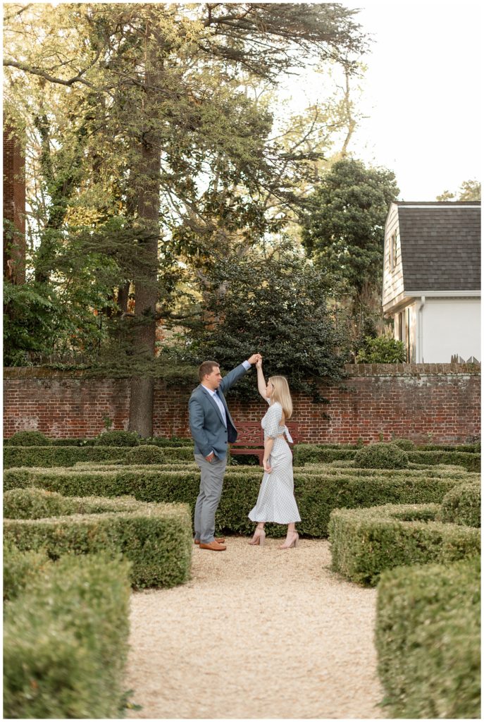 couple facing each other as guy prepares to twirl girl under his left arm in garden of manicured bushes in annapolis maryland