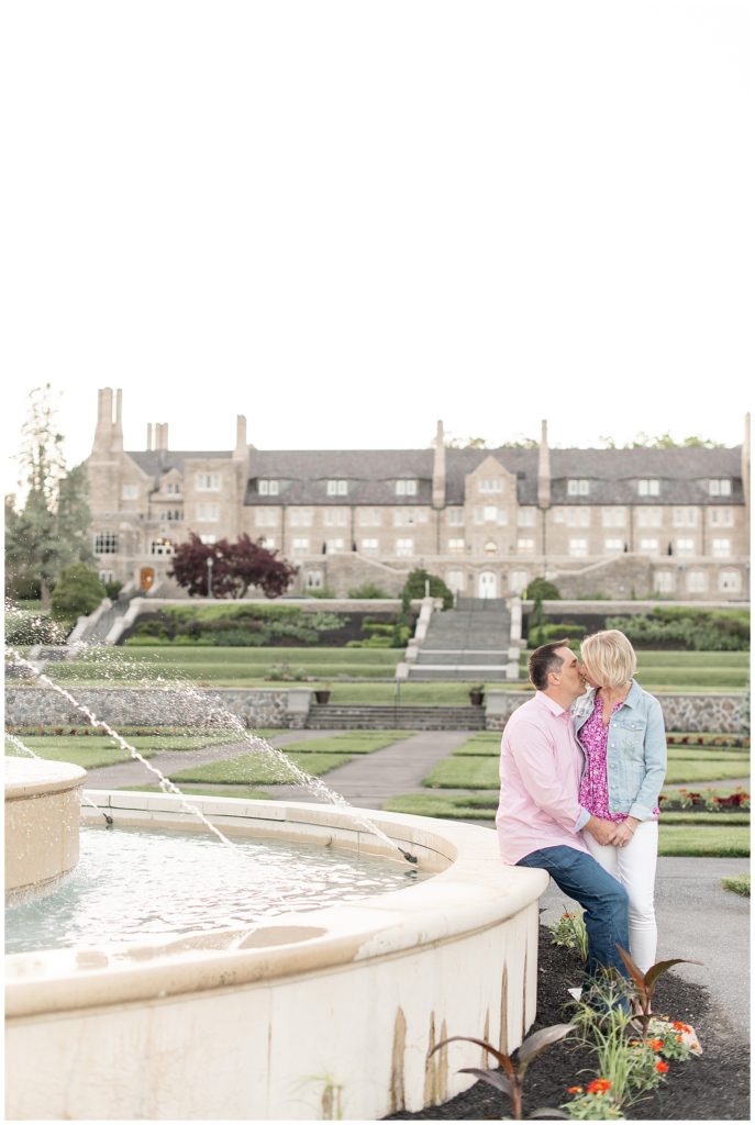 guy sitting on wall of water fountain kissing his girl with enormous building behind them at masonic village in lancaster pennsylvania