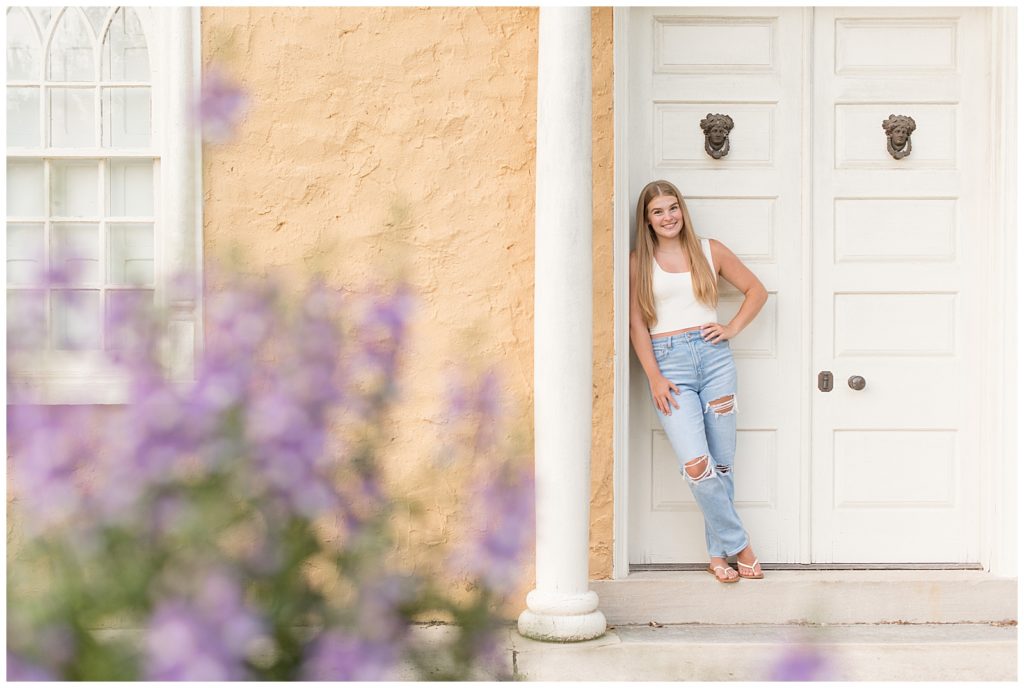 senior girl leaning against white door frame of peach colored house at hibernia park