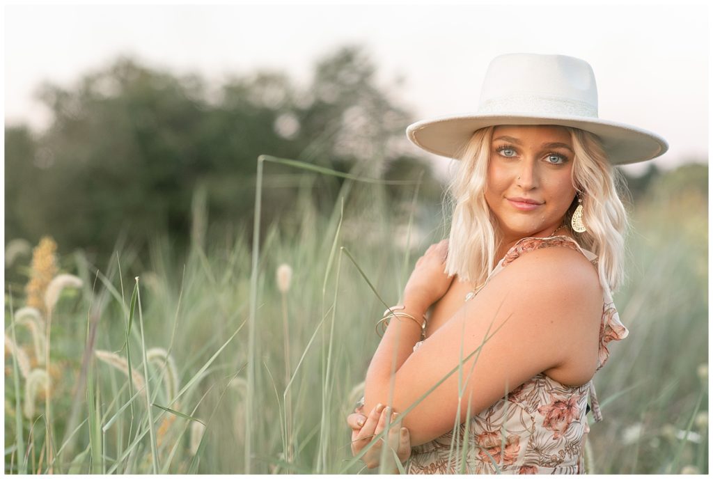 senior girl staring at camera with left hand on right shoulder wearing floral tank top and trendy white brimmed hat in tall wild grasses at overlook park