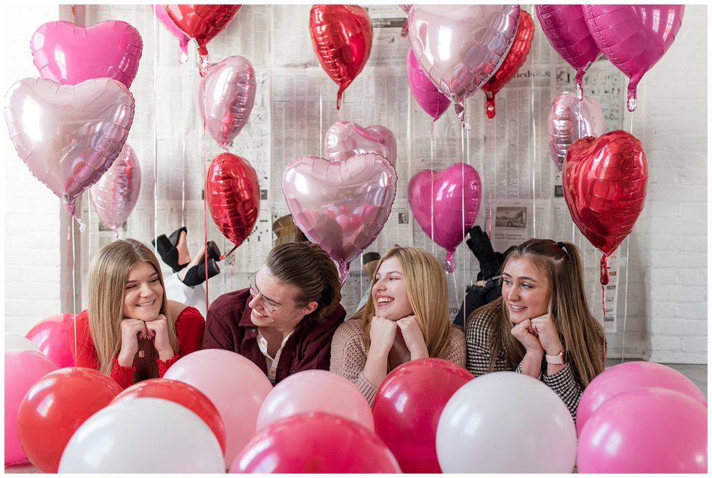 three girls and one guy laying on their stomachs surrounded by red, pink, and white balloons at hingeworks for valentine's day