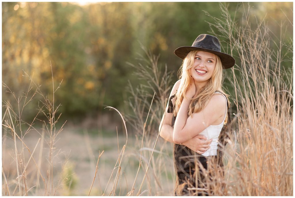 senior girl looking over left shoulder and smiling with left hand on right shoulder and wearing black overalls and trendy black hat at overlook park by tall grasses