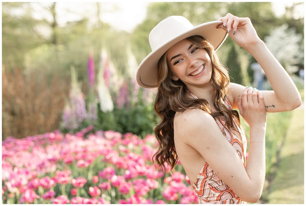 senior girl with right shoulder toward camera and head tipped back holding brim of trendy white hat by pink flowers at longwood gardens