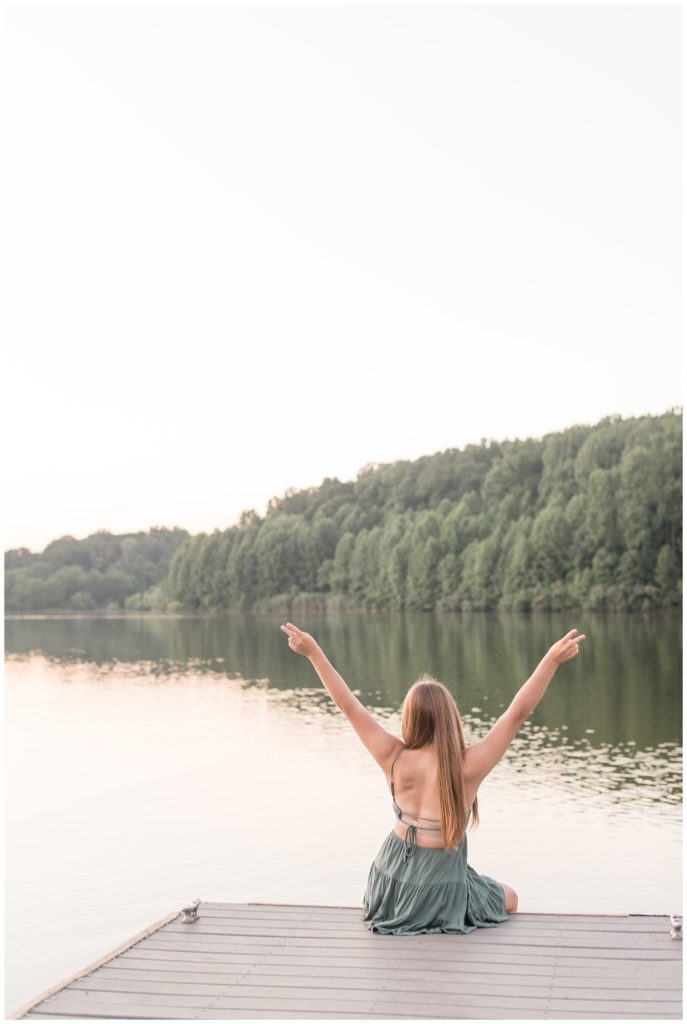 senior girl sitting on edge of dock with back towards camera and arms raised high by lake at hibernia park