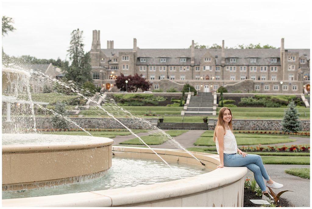 senior girl sitting on edge of concrete fountain with huge building behind her at masonic village in elizabethtown pennsylvania