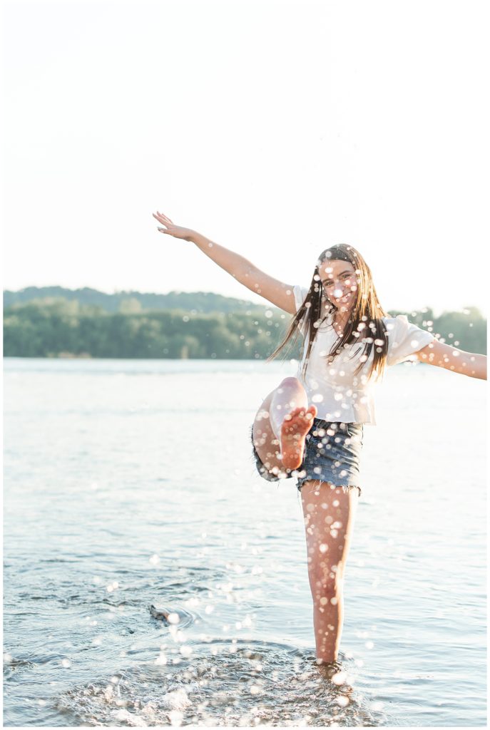 senior girl kicking her right foot towards camera and making a big splash in the susquehanna river with arms extended out on sunny evening