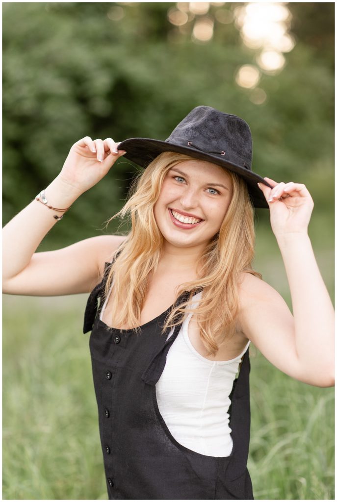 senior girl smiling at camera with both hands holding the brim of her trendy black hat at overlook park