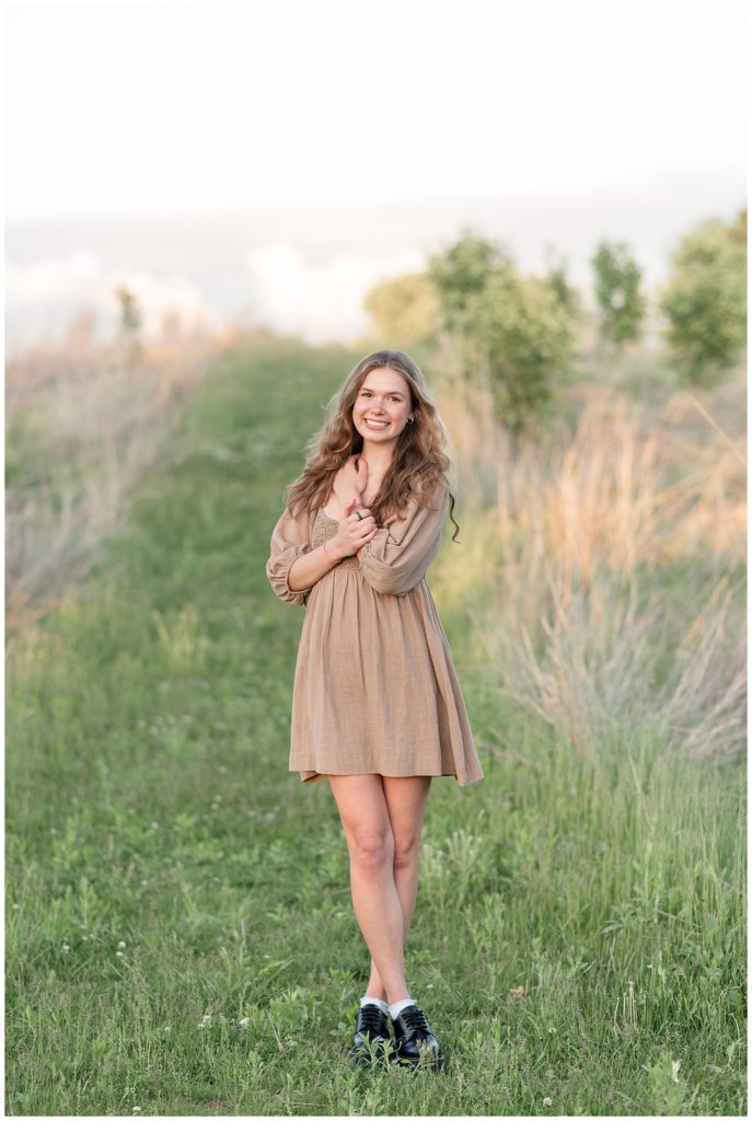 senior girl with arms crossed over her chest in tan dress among tall wild grasses at sunset at overlook park