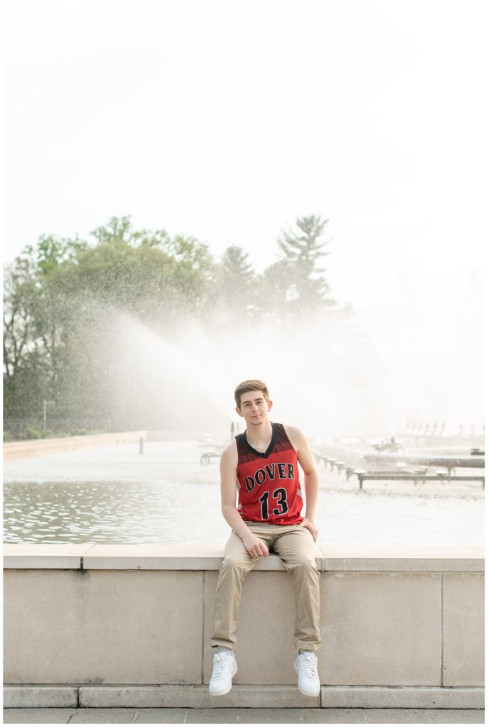 senior guy wearing red and black jersey and khaki pants sitting on ledge of fountain on sunny day at longwood gardens