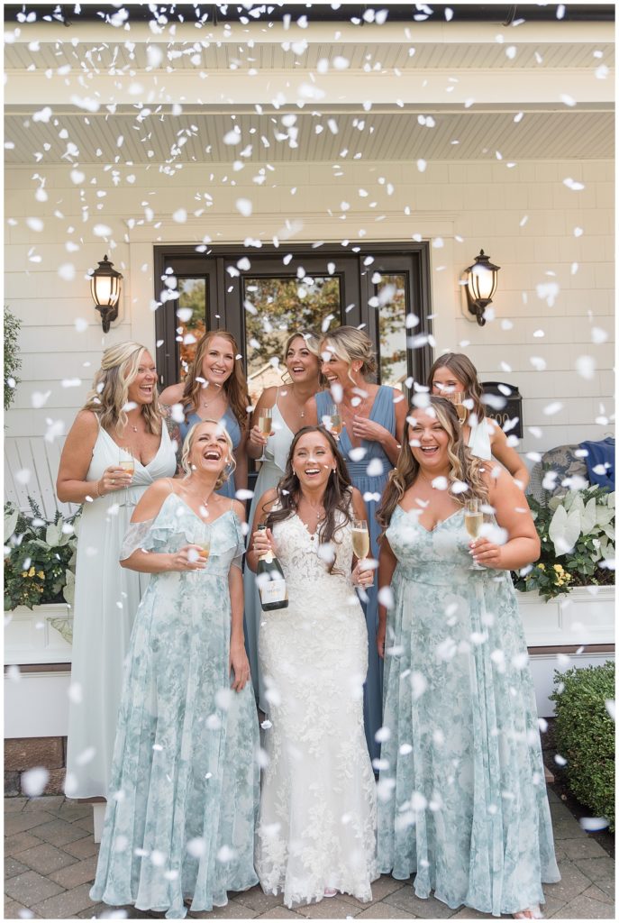 bride holding a bottle of champagne surrounded by her seven bridemaids wearing shades of blue gowns with confetti flying in new jersey