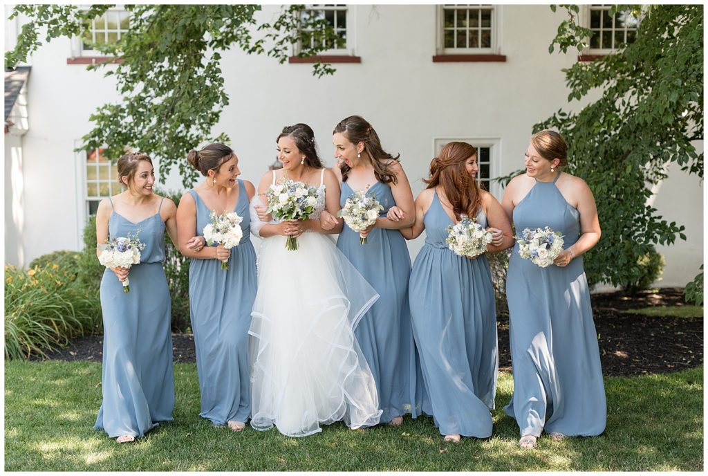 bride surrounded by her five bridesmaids wearing blue gowns and walking towards the camera smiling at each other at normandy farm
