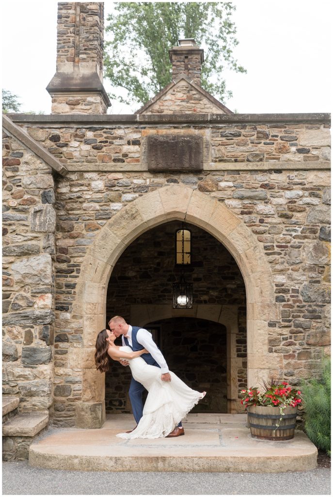 groom dipping back his bride and kissing her by beautiful stone building with archway at PARQUE Ridley Creek