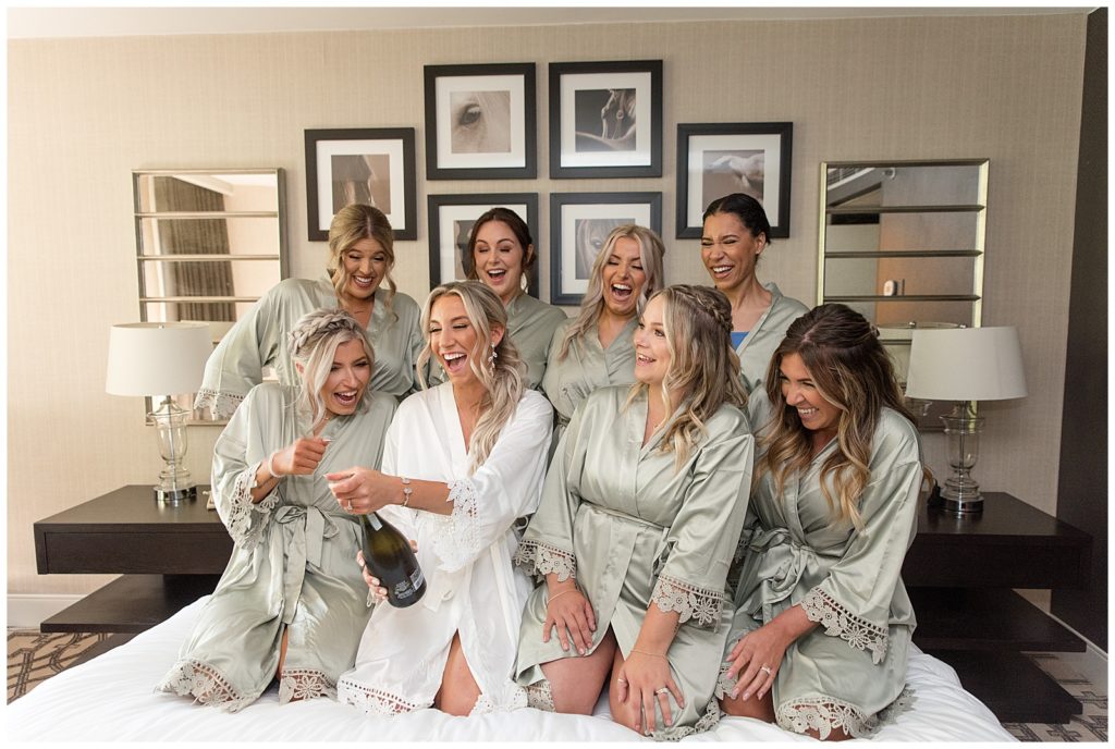 bride popping open a bottle of champagne on bed in bridal suite surrounded by her seven bridesmaids in sage robes at valley country club