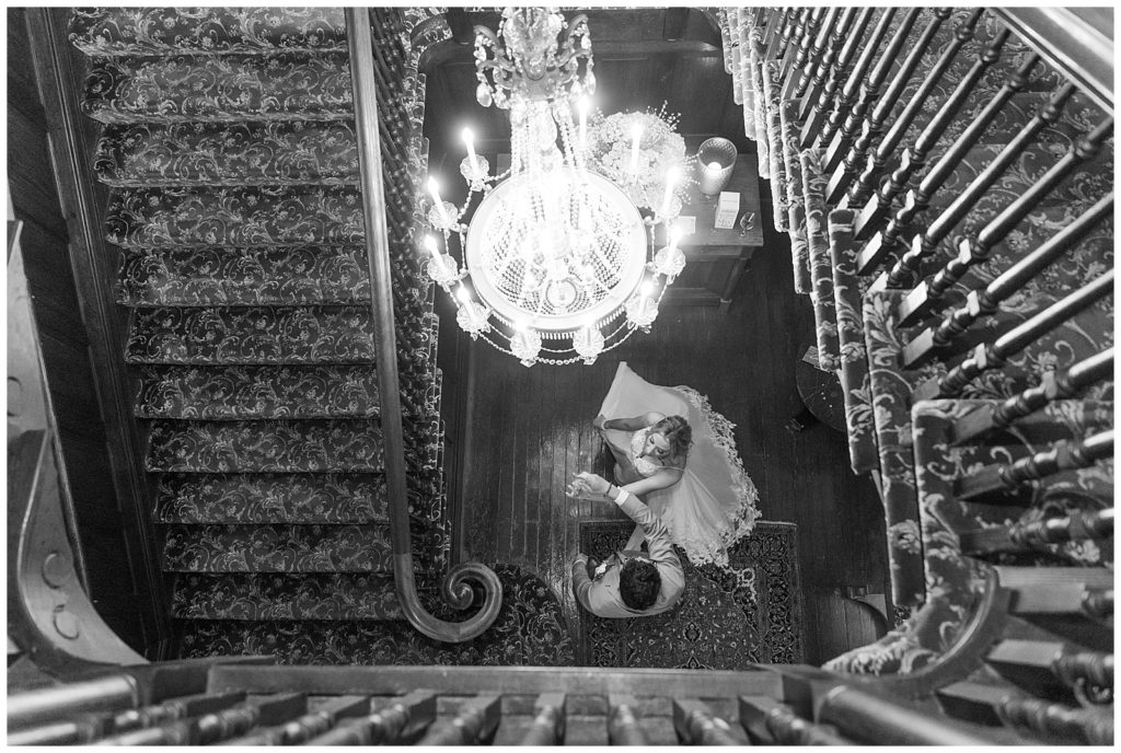 black and white photo looking down at bride and groom at bottom of spiral staircase under chandelier at the valley country club