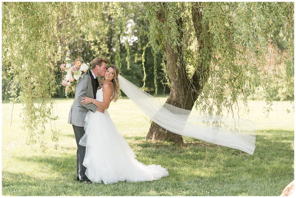 groom kissing his bride's right cheek as her long veil blows behind her by willow tree at the barn at silverstone