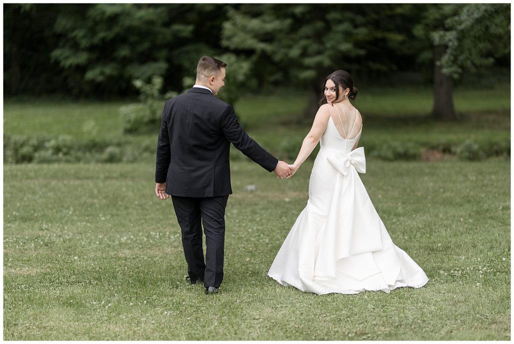 couple holding hands and walking away from camera as bride looks back in grass field at cameron estate inn in lancaster county