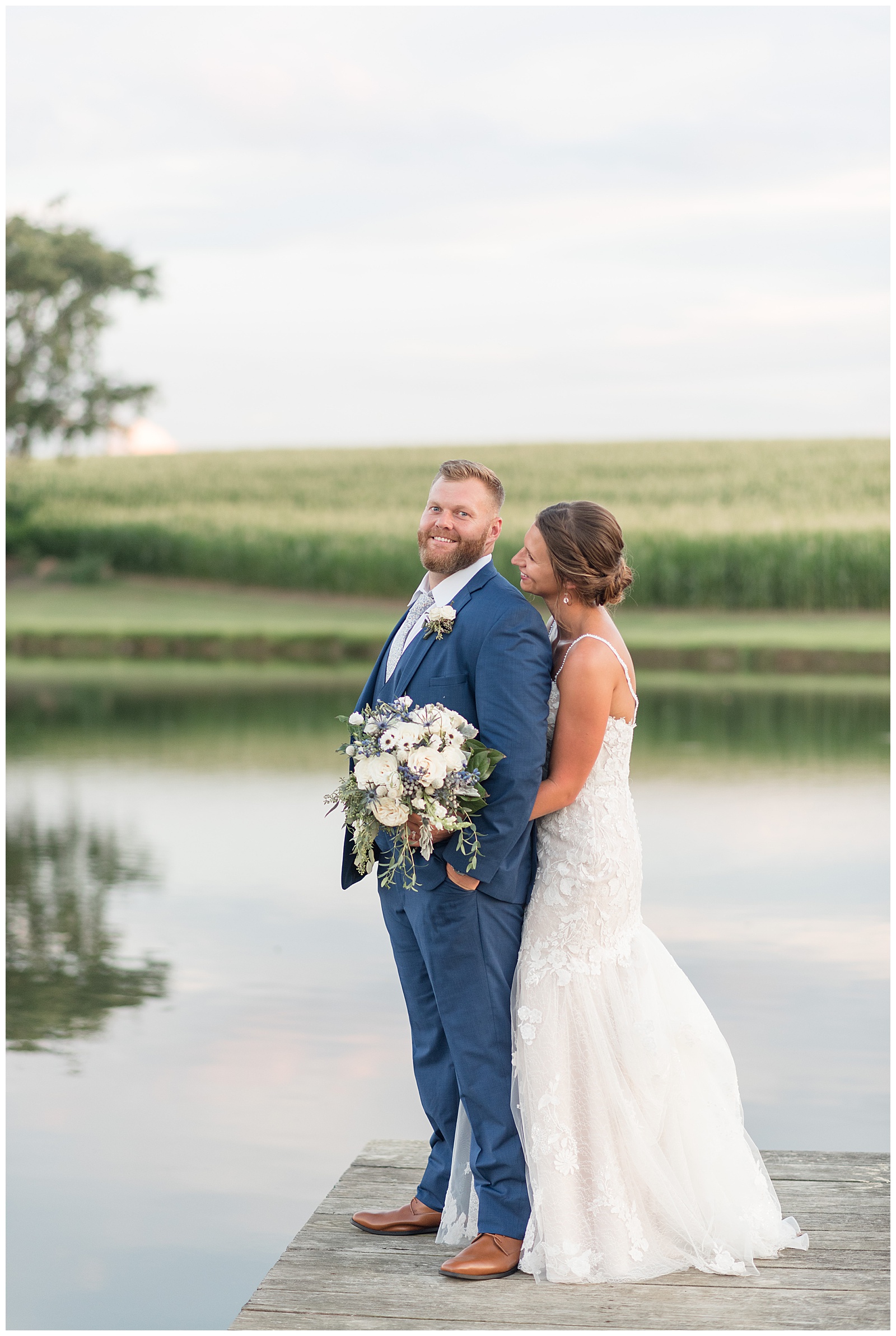 bride standing behind her groom on dock by pond at lakefield weddings with cornfield in background