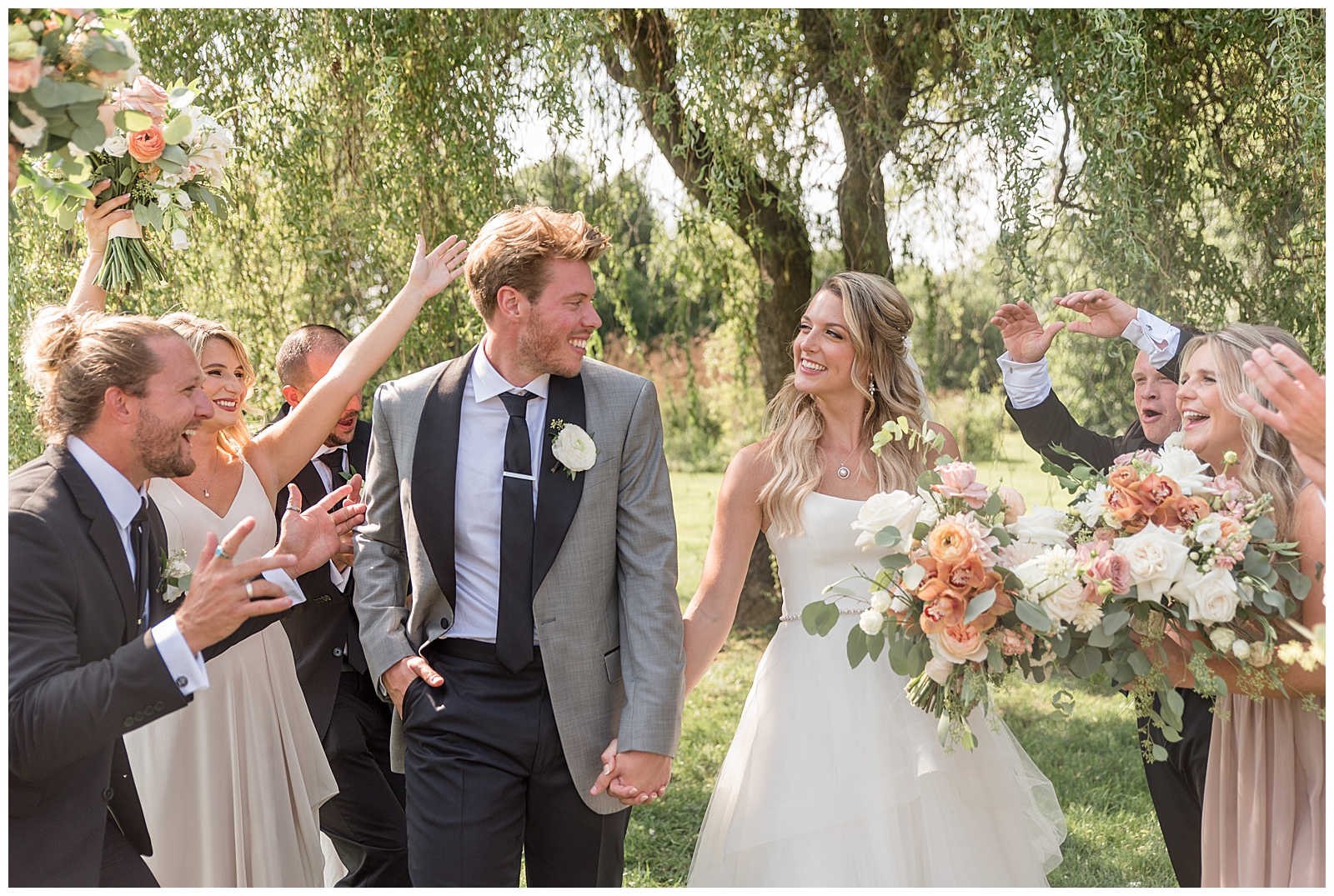 bride and groom smiling and holding hands as they walk between two rows of their bridal party by willow tree at the barn at silverstone