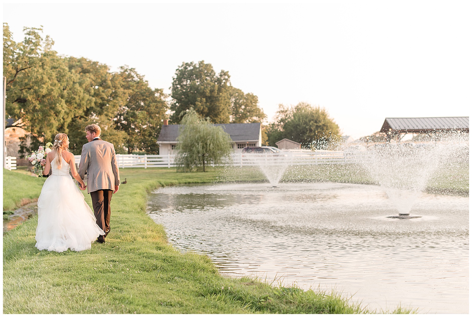couple walking around pond with fountains with their backs toward the camera on sunny evening at the barn at silverstone in lancaster county