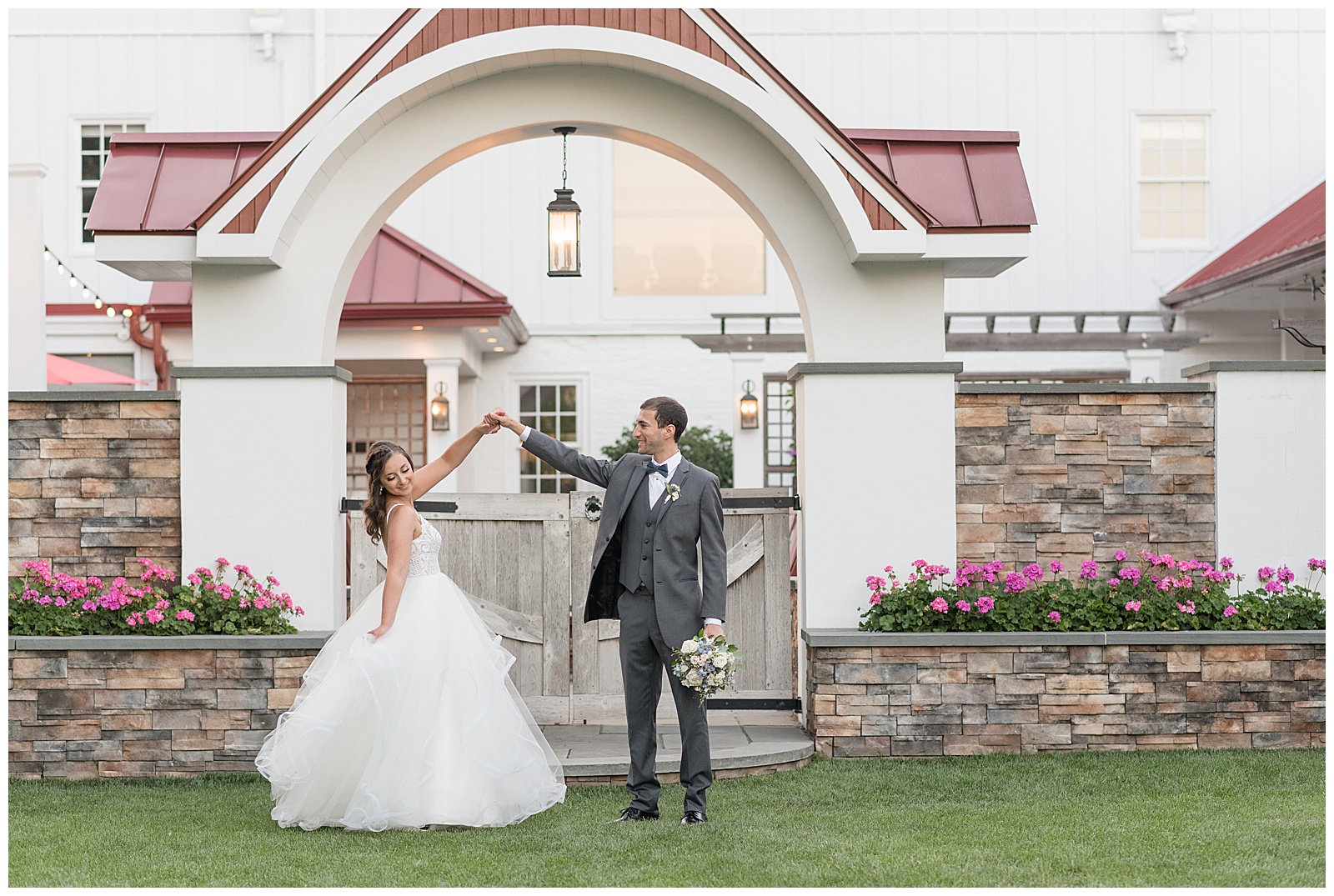 groom twirling his bride under his right around as she holds her dress train out under large archway at normandy farm