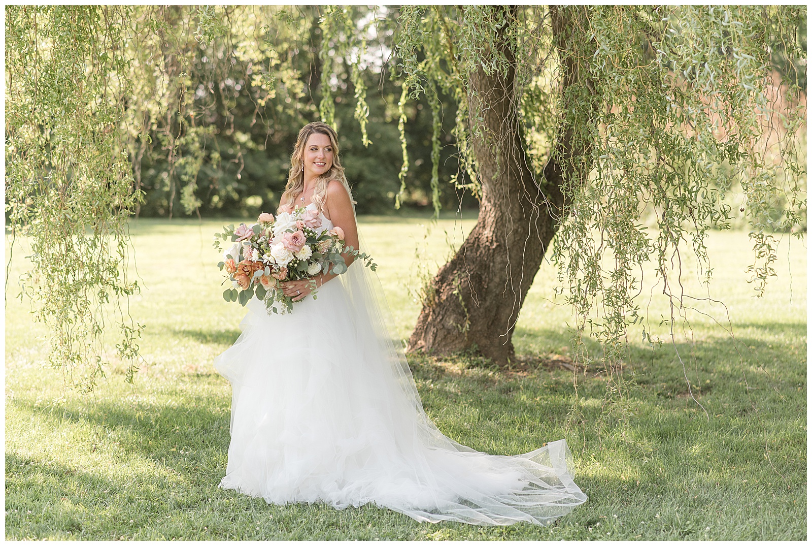 bride standing under willow tree holding stunning bouquet on sunny day at the barn at silverstone