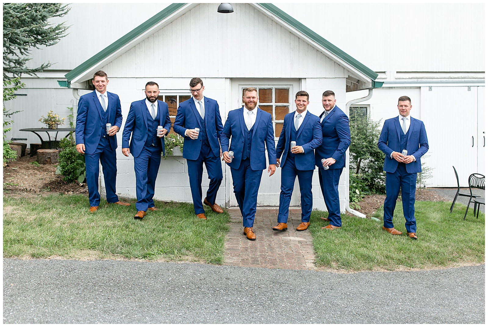 groom with his six groomsmen all wearing navy suits walking towards camera by white barn at lakefield weddings