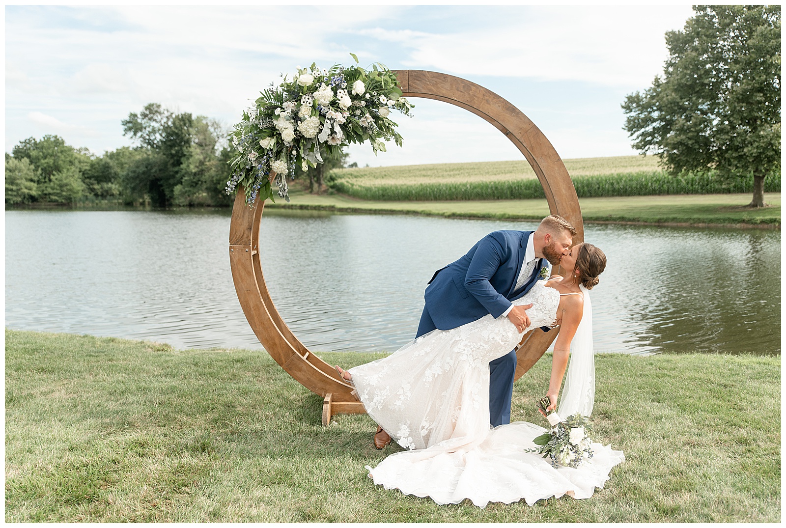 groom dipping his bride back as they kiss by wooden and floral structure by pond at lakefield weddings