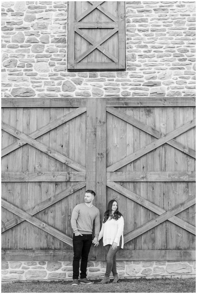 black and white photo of engaged couple holding hands and looking in opposite directions by huge wooden door at folino estate winery