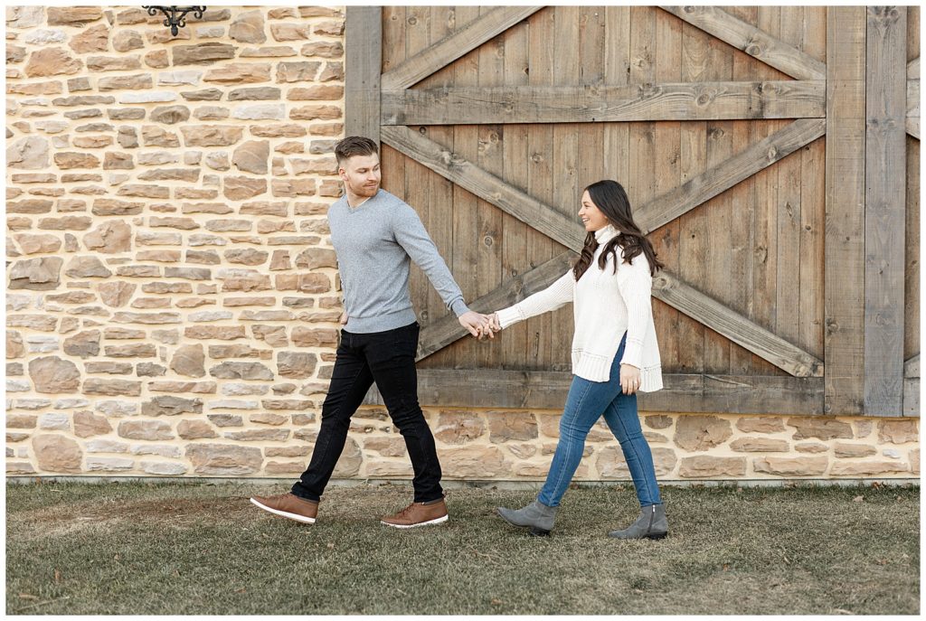 guy holding girl's hand and leading her as he looks back at her by wooden door and stone building at winery in kutztown pennsylvania