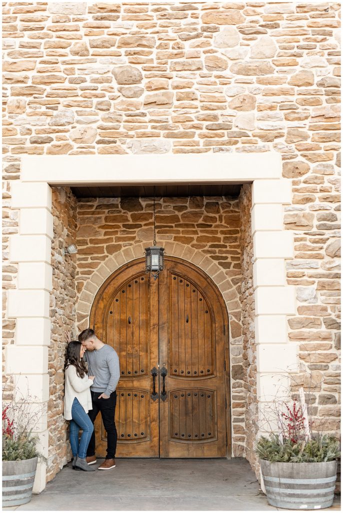 girl leaning against stone door frame by huge wooden door as her fiance hugs and rests his forehead against her at folino estate