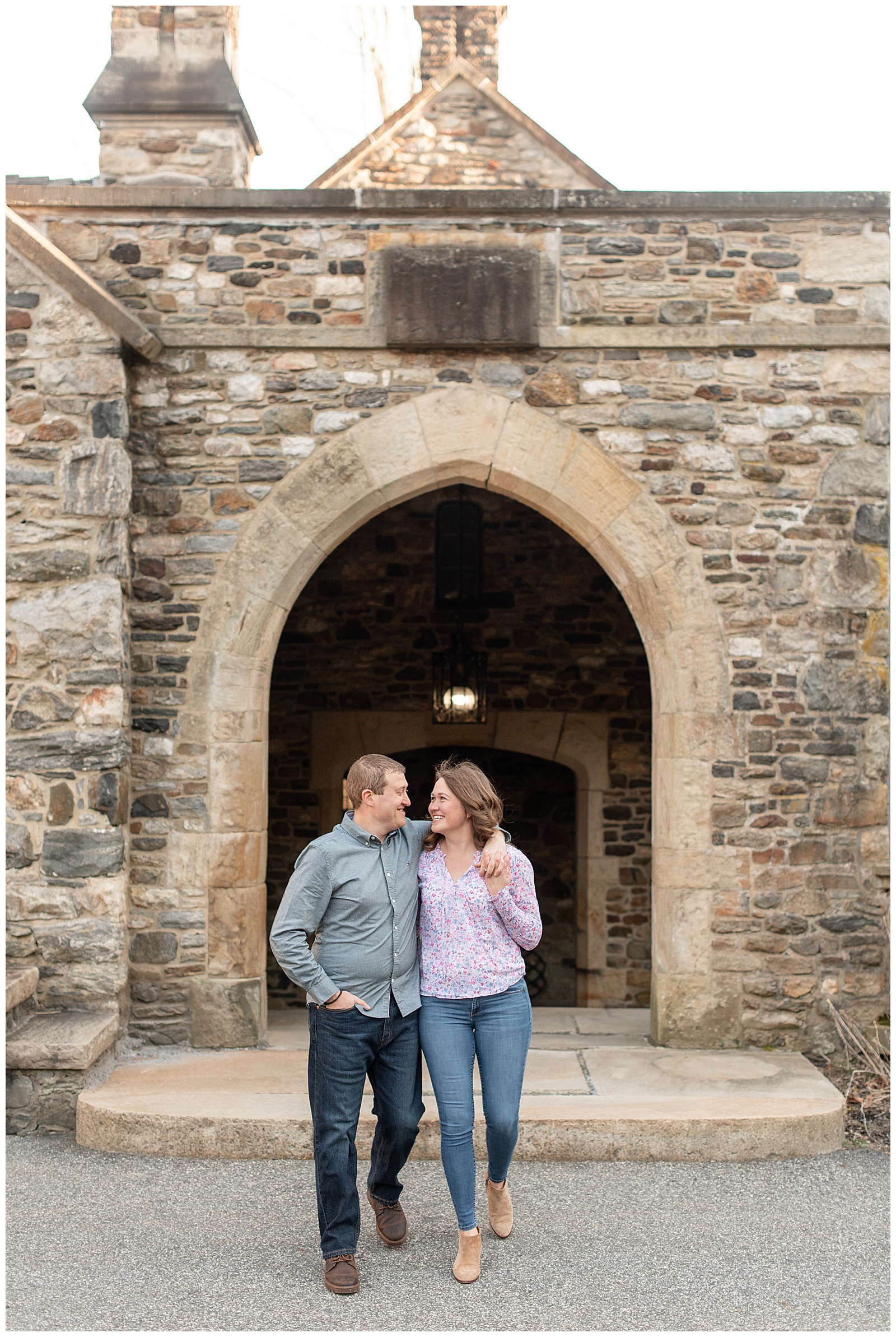 engaged couple standing by stone archway looking at each other smiling on winter day at ridley creek park