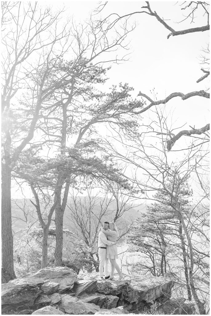 black and white photo of engaged couple hugging and kissing with trees behind them at pinnacle point overlook 