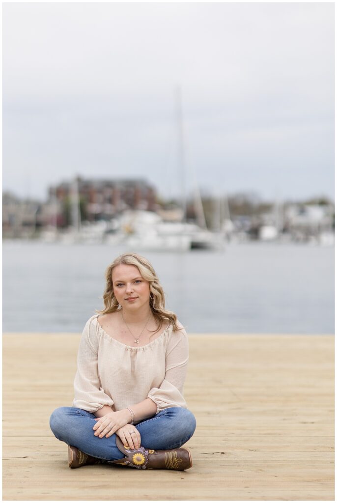 college senior girl sitting on dock with bay behind her in beige top and blue jeans in annapolis maryland