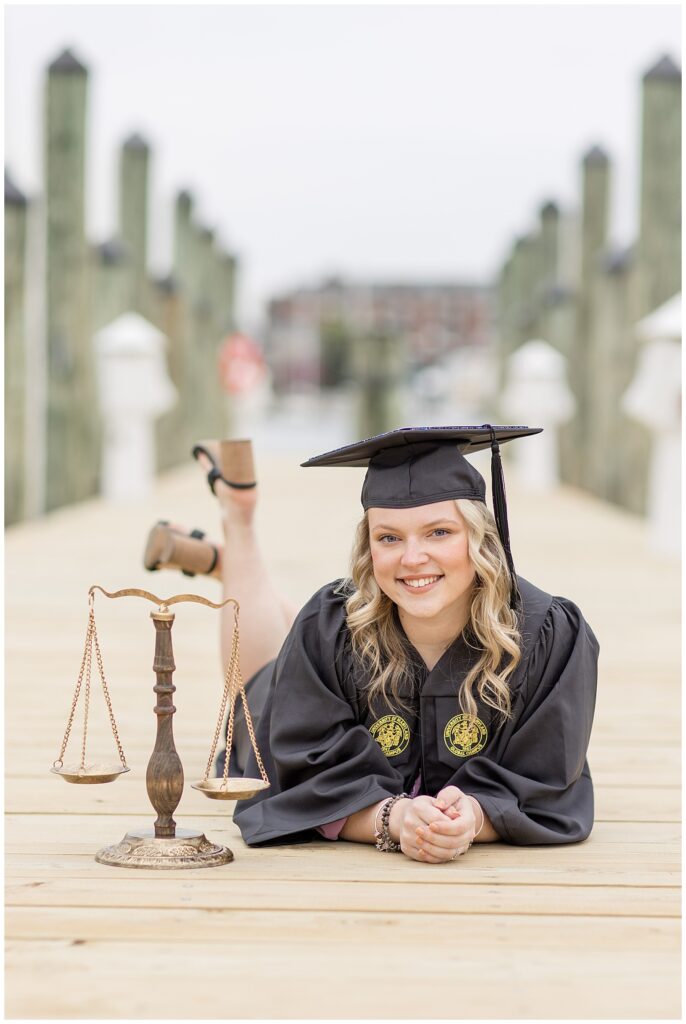 college senior girl wearing black cap and gown laying on stomach on wooden dock resting on her forearms in maryland