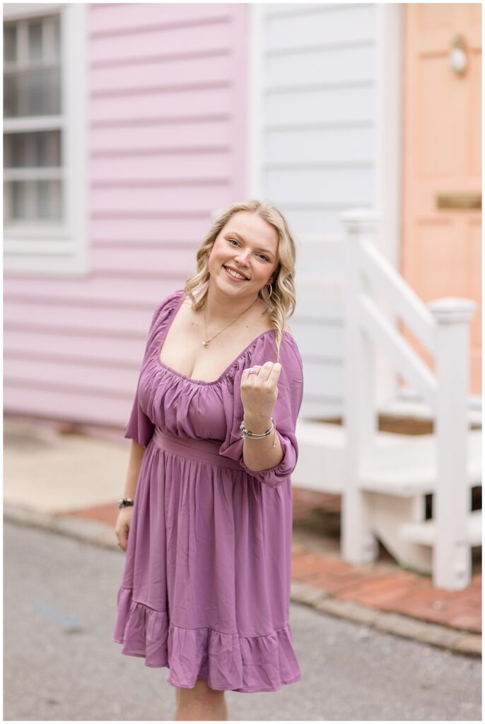 college senior girl wearing purple dress standing with house behind her and her left hand holding one of her long curls in annapolis