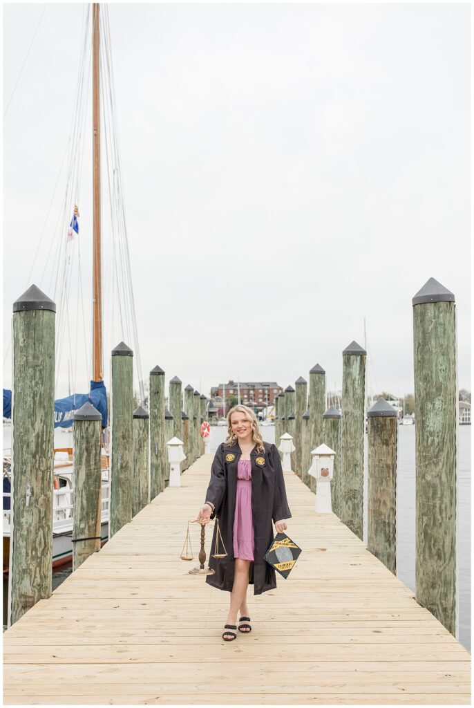senior girl wearing opened up black graduation gown with purple dress showing on wooden dock in annapolis maryland