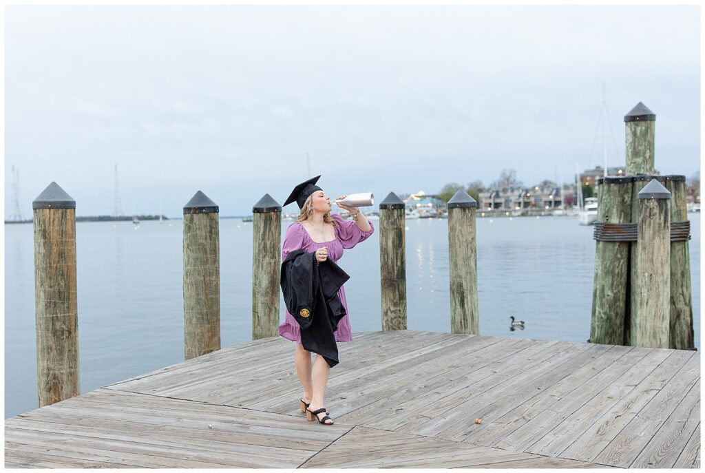 college senior girl holding black graduation gown and wearing black graduation cap taking a drink on dock in annapolis maryland