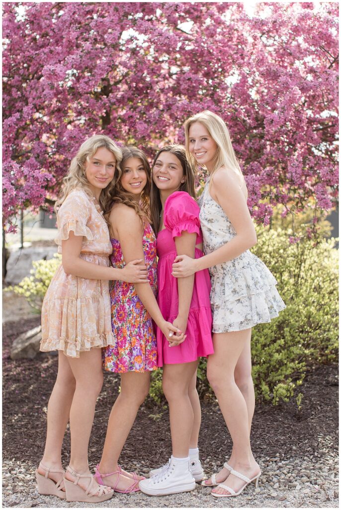 four senior girls wearing cute short dresses in shades of pink by pink tree at overlook park huddled together