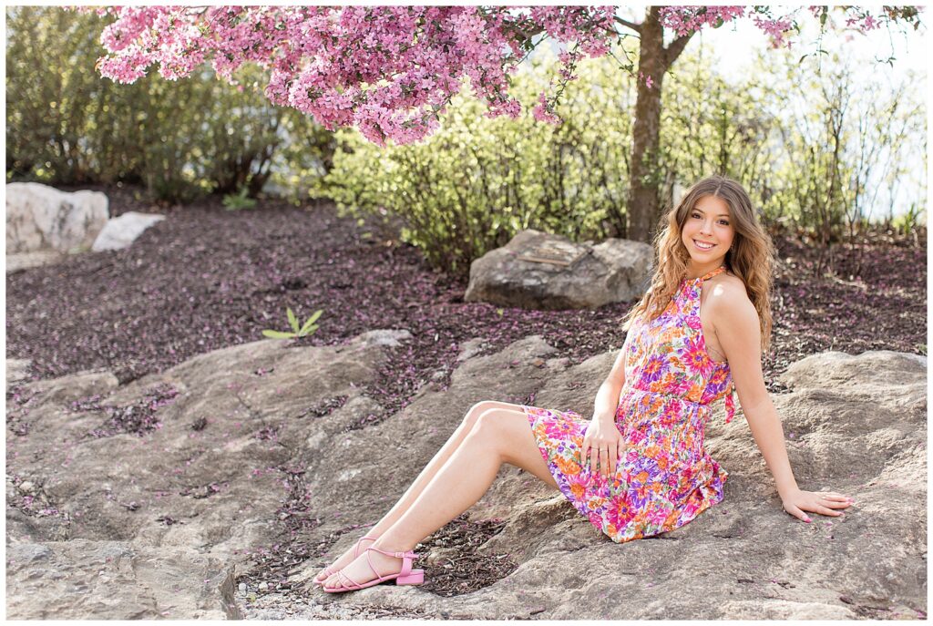 senior girl with bright pink floral dress leaning back as she sits on large rock at overlook park