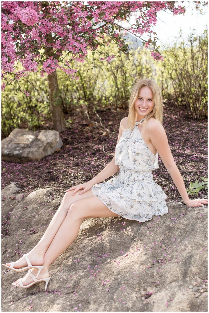senior girl wearing white floral dress sitting on large rock and smiling on spring evening at overlook park