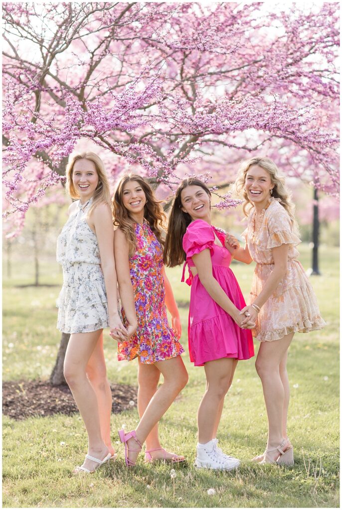 four senior girls facing different directions and holding hands and smiling by pink tree at manheim township park