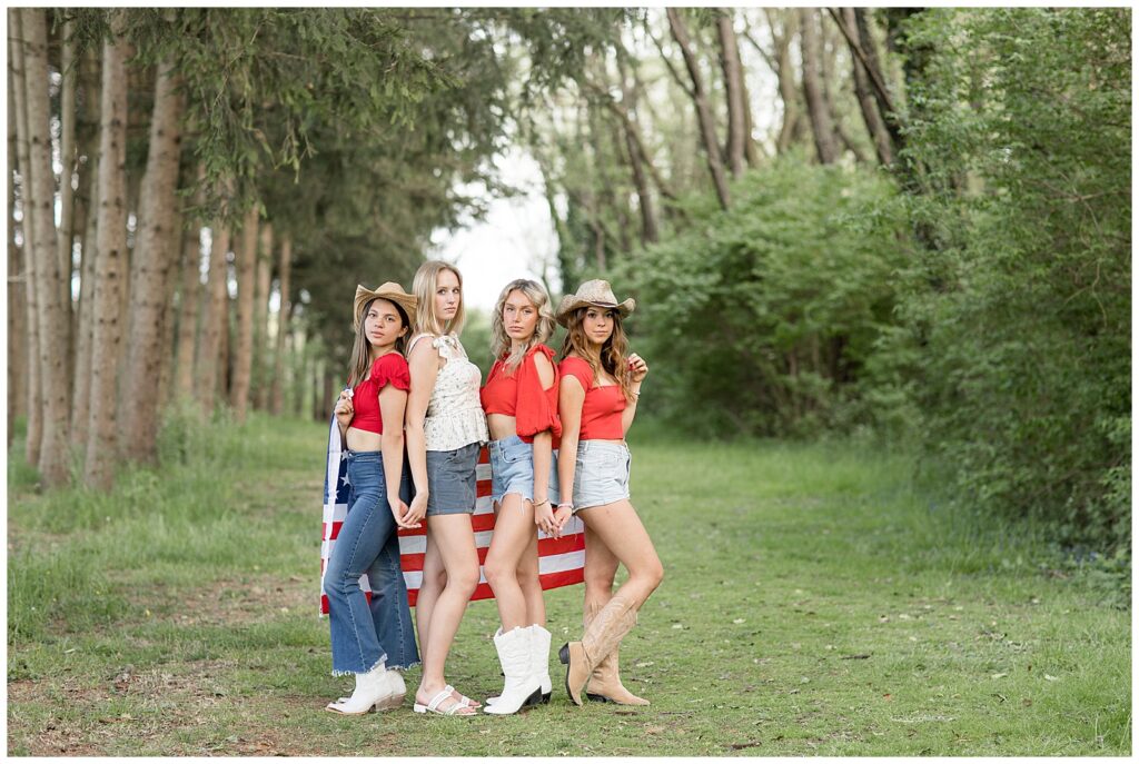 four senior girls wearing red, white, and blue standing by American flag and posing at park in lancaster county