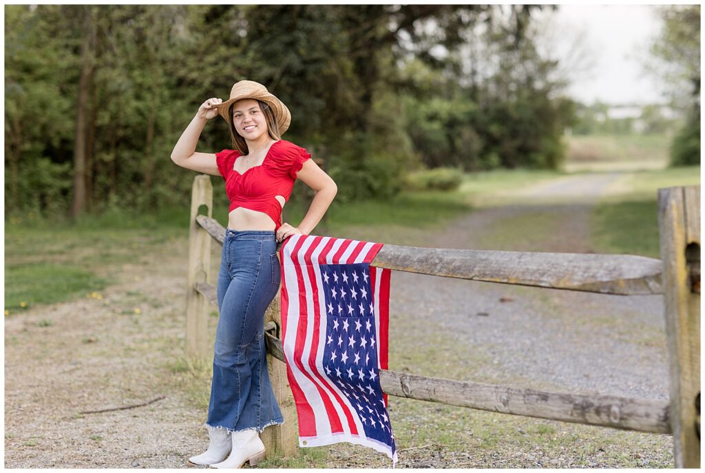 Senior girl wearing red shirt, blue jeans, and cowboy hat leaning against fence by american flag at overlook park